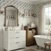 Blue floral ceramic tile adding vintage cottagecore charm to a bright bathroom with a white fluted vanity, brass fixtures, classic beadboard, and botanical décor.