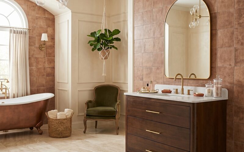 Walnut wood bathroom vanity with rectangular gold mirror and brass sconces against full room terracotta square ceramic wall tile in warm brown tones, stone tile flooring with vintage rug and crystal chandelier